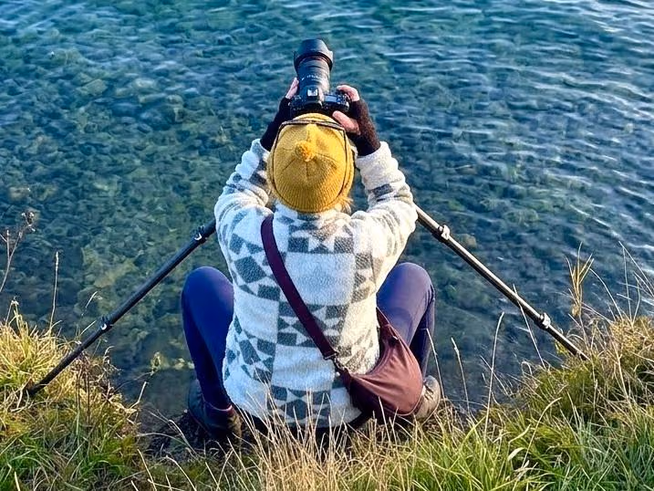 Janet von hinten, im Gras sitzend mit Stativ am klaren Wasser